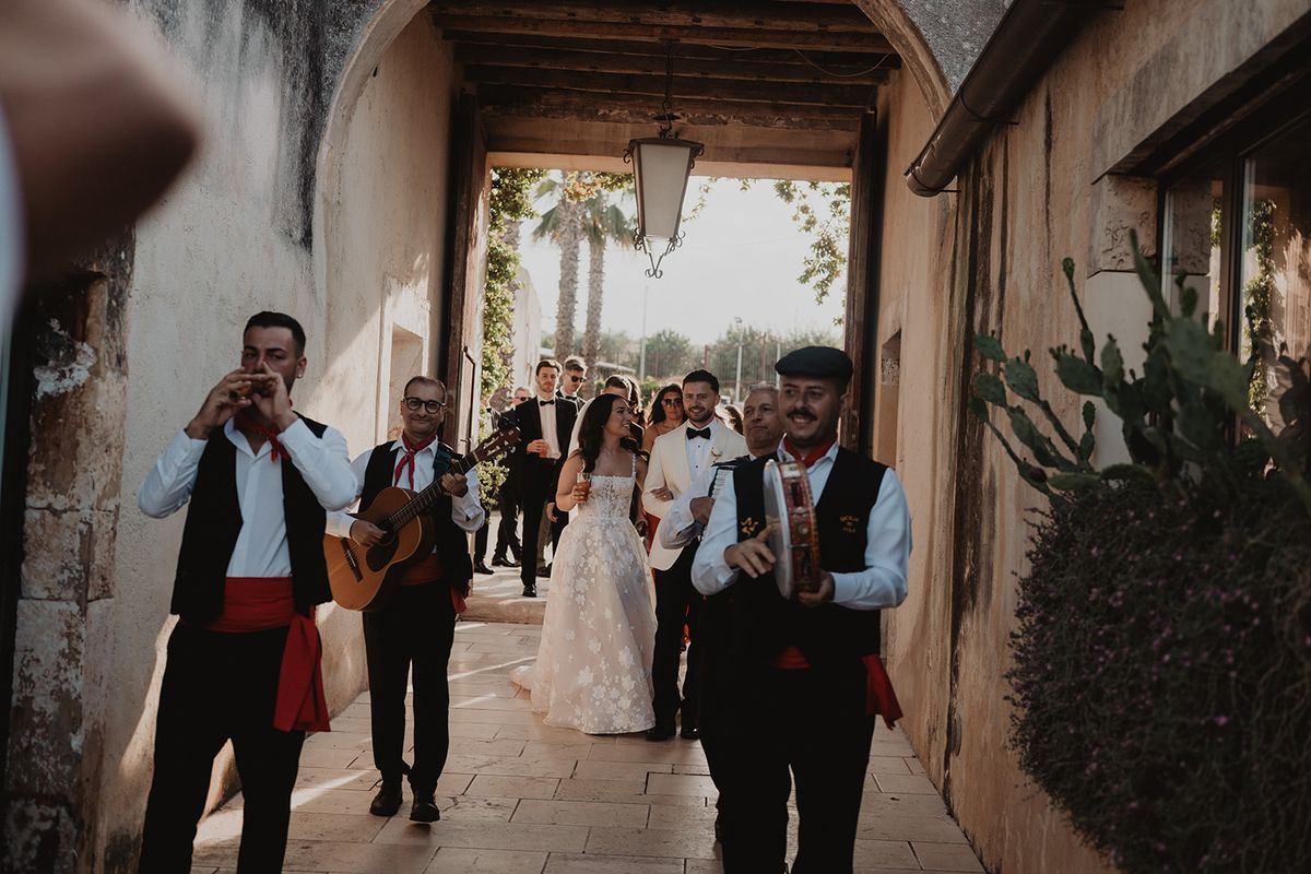 Newlyweds led by traditional Sicilian folk musicians through a honey-coloured stone archway