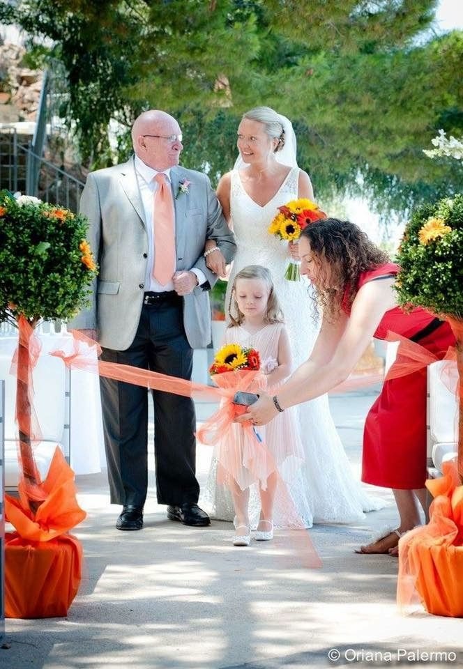 Lina Benfeito in a red dress untying the orange aisle ribbon as the bride, her father and the flower girl wait to walk down the aisle at a Sicilian wedding ceremony